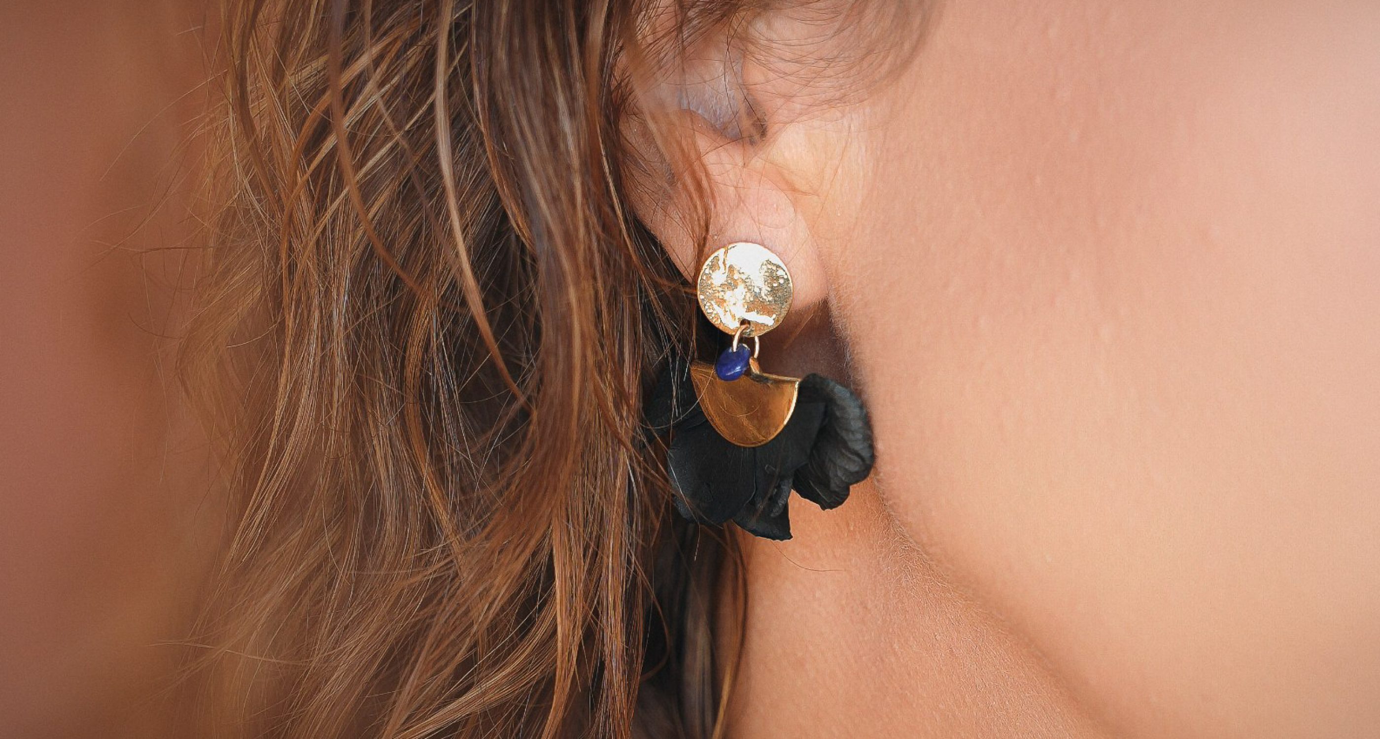 Minuit studio brand photography – close up on a high end earring jewelry made of gold and black flower petals on a brunette person with a brown caramel brackground for a French flair to fashion visuals Minuit studio brand photography - close up on a high end earring jewelry made of gold and black flower petals on a brunette person with a brown caramel brackground for a French flair to fashion visuals